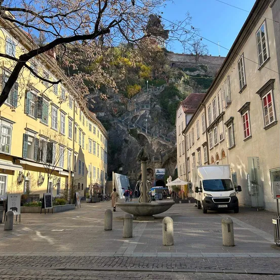 Eine Straßenszene mit einem Steinbrunnen in der Mitte, einem gelben Gebäude auf der linken Seite, einem weißen Lieferwagen auf der rechten Seite und einem großen Steinfelsen im Hintergrund.
