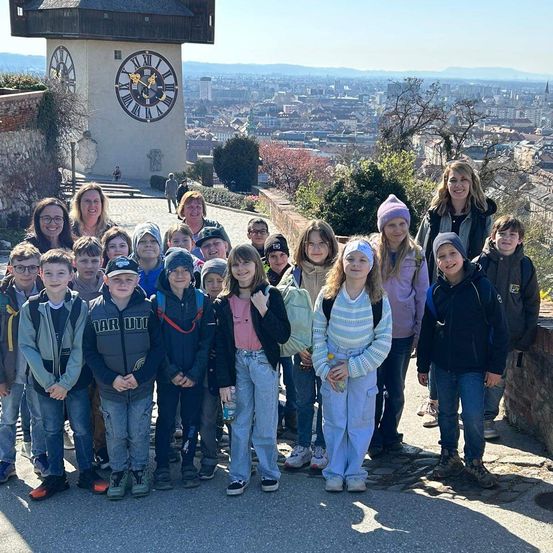 Eine Gruppe von Kindern und ihrer Lehrerin steht auf einem Weg vor einem großen Uhrturm. Der Turm befindet sich inmitten einer Stadt mit vielen Gebäuden und Bäumen.