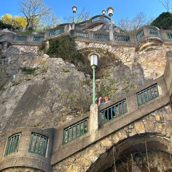 Eine Frau lehnt sich an ein Geländer mit grünen Metallstäben auf einer Steinbrücke, die an einem sonnigen Tag über einer Felswand verläuft.