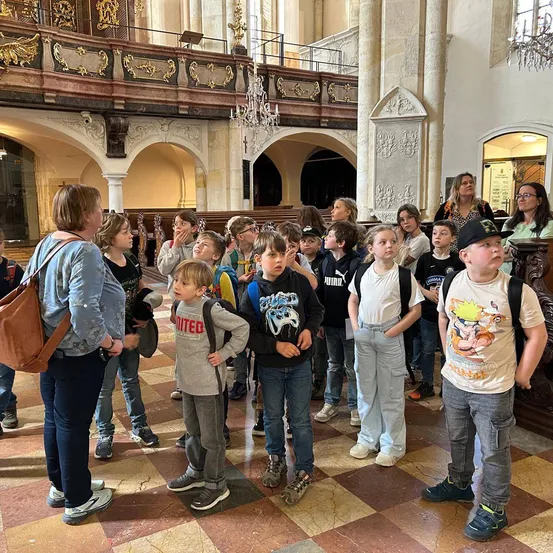 Eine Gruppe von Kindern und zwei Erwachsenen steht im Inneren einer Kirche. Die Kinder tragen Ruckscke und Turnschuhe.