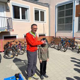 Ein Mann in einem roten Jacket und Brille hält den Helm eines Kindes vor einem Gebäude, vor dem Fahrräder geparkt sind.