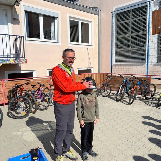 Ein Mann in einem roten Jacket und Brille hält den Helm eines Kindes vor einem Gebäude, vor dem Fahrräder geparkt sind.