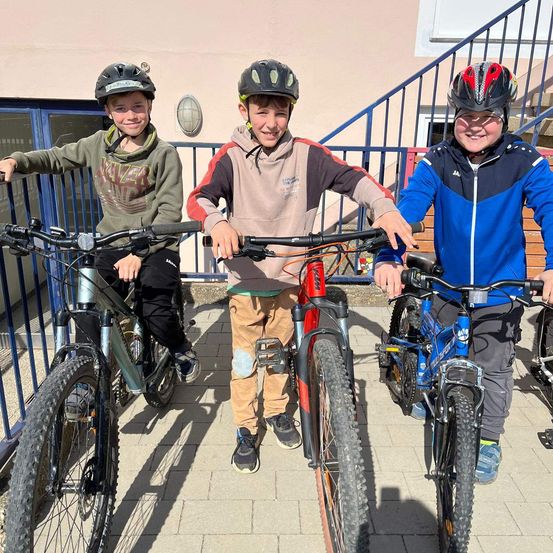 Drei junge Jungen mit Helmen posieren für ein Foto mit ihren Fahrrädern. Sie lächeln und scheinen viel Spaß zu haben. Sie stehen vor einem Gebäude mit einer Treppe.