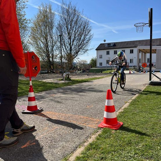 Ein Kind fährt mit dem Fahrrad auf einem Schulparkplatz, während eine Person in Rot in der Nähe geht.