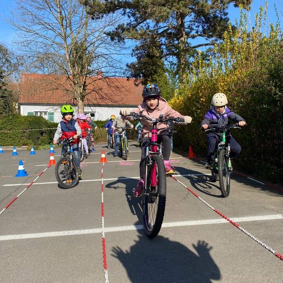Mehrere Kinder fahren auf einer markierten Strecke mit orangefarbenen und blauen Hütchen auf Fahrrädern, alle tragen Helme. Sie befinden sich vor einem Haus mit rotem Dach.