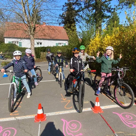 Mehrere Kinder fahren mit Fahrrädern auf einem Parkplatz. Sie tragen alle Helme. Ein Haus mit rotem Dach ist im Hintergrund.