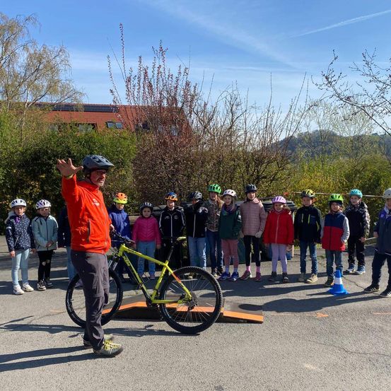 Ein Mann in einer orangefarbenen Jacke winkt einer Gruppe von Kindern zu, die Helme tragen. Sie stehen hinter einem gelben Fahrrad auf einer Rampe, mit einem orangefarbenen Kegel an der Seite. Dahinter befindet sich ein Gebäude mit Solarmodulen und einem Bergrücken.