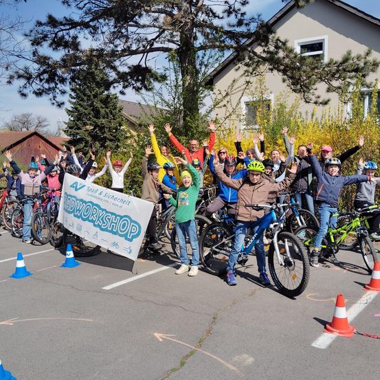 Gruppe von Menschen, Erwachsene und Kinder, mit Helmen, stehen in einem Parkplatz mit Fahrrädern. Ein Banner vor ihnen sagt 'Roadworkshop'.