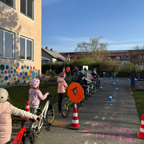 Eine Gruppe von Kindern mit Fahrrädern, die Helme tragen, steht in einer Schule an einer Reihe, mit einem Stoppschild in der Mitte, umgeben von orangefarbenen Kegel und einer bunten Wand an der Seite.