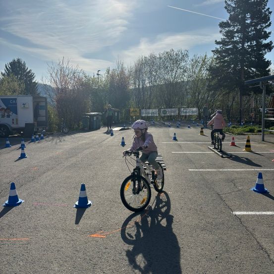 Zwei Kinder fahren auf einem Parkplatz mit ihren Fahrrädern um Verkehrskegel herum. Eine Person steht in der Nähe und beobachtet. Im Hintergrund befinden sich ein LKW, Bäume und ein klarer Himmel.