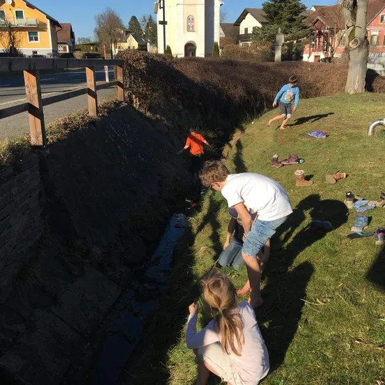 Drei Kinder spielen in einem kleinen Bach in einem Grasbereich, mit Häusern und einer Kirche im Hintergrund.