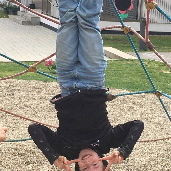 Ein junges Kind hängt kopfüber von einer Kletterstruktur. Es trägt ein schwarzes Shirt und blaue Jeans.