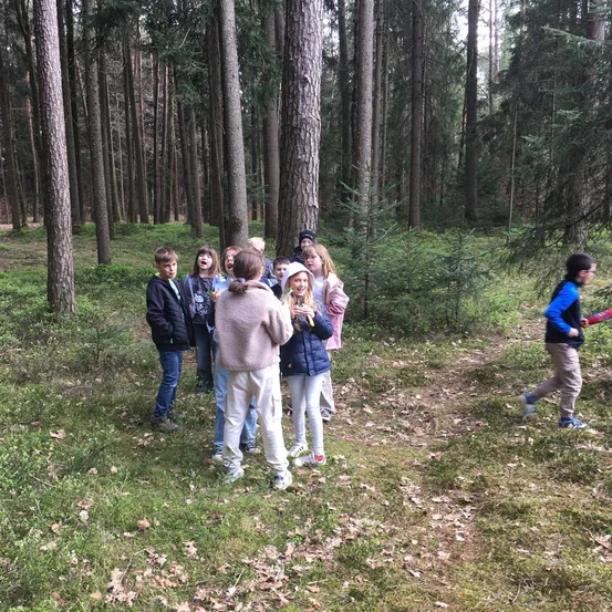 Eine Gruppe von Kindern steht in einem Wald, wobei eines auf sie zuläuft. Sie tragen alle Jacken, Hosen und Turnschuhe. Einige von ihnen tragen Mützen.