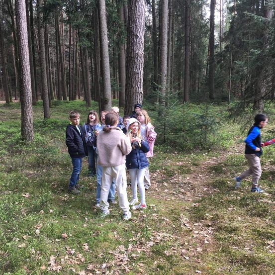Eine Gruppe von Kindern steht in einem Wald, wobei eines auf sie zuläuft. Sie tragen alle Jacken, Hosen und Turnschuhe. Einige von ihnen tragen Mützen.