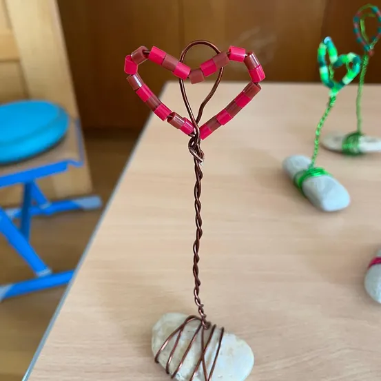 Ein herzförmiges Handwerk aus Kupferdraht und rosa Perlen steht auf einem Holztisch. In der Nähe sind weitere Kunstwerke, darunter ein grün umwickeltes Herz.