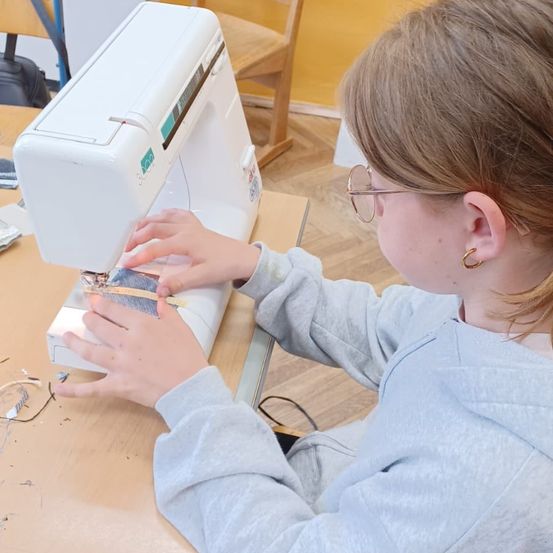Ein junges Mädchen benutzt eine weiße Nähmaschine auf einem Holztisch, trägt eine Brille und eine graue Kapuzenjacke. Die Maschine hat ein Stück Stoff, das genäht wird.