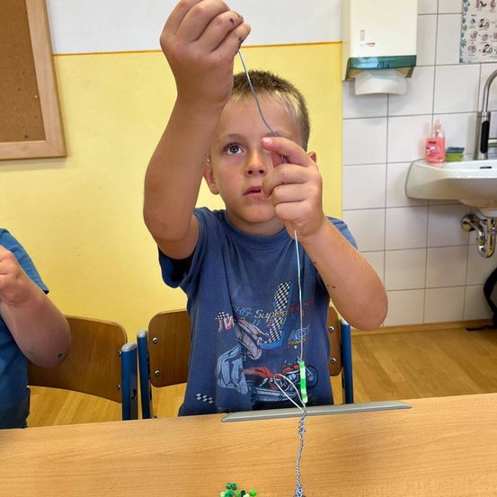 Ein junger Junge sitzt an einem Schreibtisch und hält eine Schnur mit Perlen, möglicherweise in einer schulischen Aktivität. Ein Waschbecken und ein Papierhandtuchspender stehen hinter ihm.