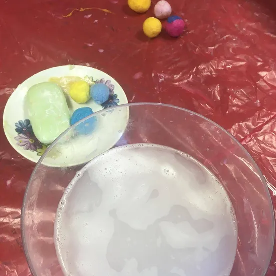 Ein Glas mit Seifenwasser steht auf einem roten Tisch neben einem Teller mit Seife und bunten Bällen.