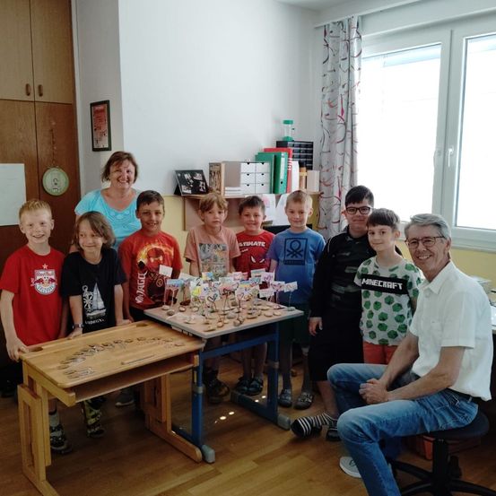 Eine Gruppe von Kindern und zwei Erwachsenen steht um einen Holztisch mit einem Bastelprojekt in einem Raum.