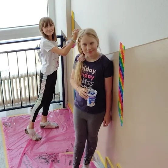 Zwei junge Mädchen malen eine Wand. Eine steht auf einem rosa Tuch und hält einen Pinsel, während die andere einen Farbbecher in der Hand hat. Beide haben ein Lächeln im Gesicht.