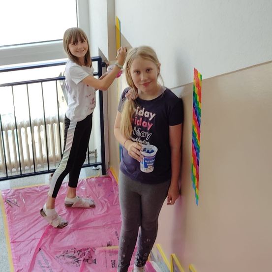 Zwei junge Mädchen malen eine Wand. Eine steht auf einem rosa Tuch und hält einen Pinsel, während die andere einen Farbbecher in der Hand hat. Beide haben ein Lächeln im Gesicht.