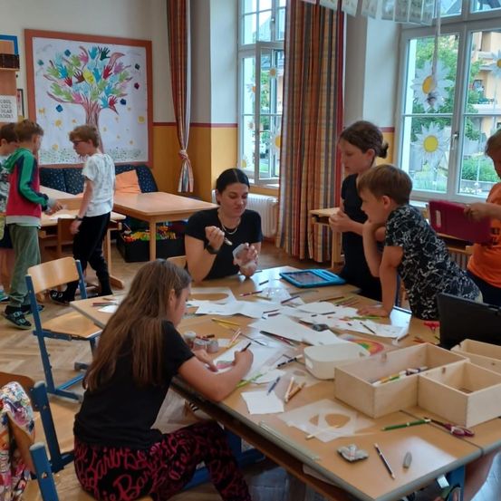 Kinder arbeiten in einem Klassenzimmer mit Fenstern und einer Wandmalerei an einem Tisch mit Malutensilien.
