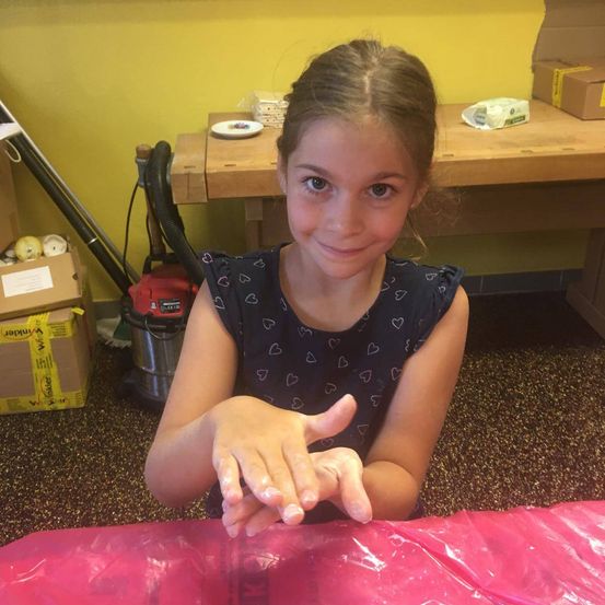 Ein junges Mädchen lächelt in die Kamera, ihre Hände sind mit einer weißen Substanz bedeckt. Sie sitzt an einem Tisch mit einer pinken Plastikfolie, einem gelben Karton und einem Staubsauger hinter ihr.