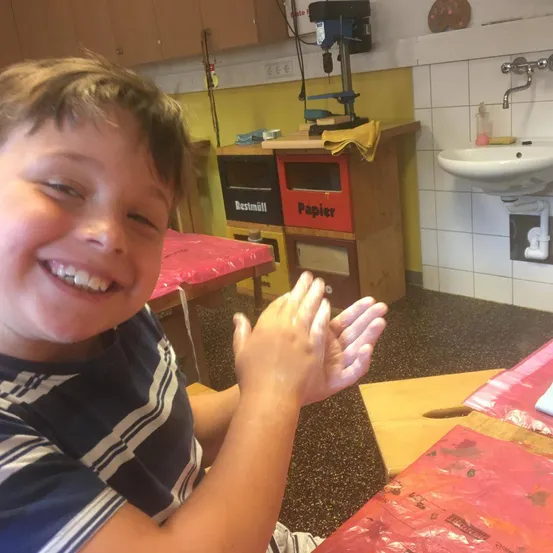 Ein junger Junge sitzt mit erhobenen Händen an einem Tisch. Er lächelt und scheint sich in einem Klassenzimmer zu befinden. Hinter ihm steht ein Tisch mit einer roten Abdeckung. An der Wand befinden sich Schränke und eine Steckdose.
