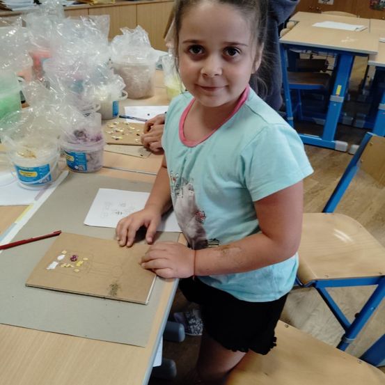 Ein junges Mädchen steht in einem Klassenzimmer, lächelt und hält ein Stück Papier mit bunten Steinen. Hinter ihr stehen verschiedene Plastiktüten und Behälter auf einem Tisch.