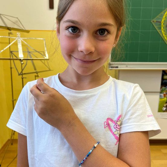 Ein junges Mädchen mit blonden Haaren lächelt und trägt ein weißes T-Shirt mit einer rosa Cartoon-Figur und einem Armband. Sie befindet sich in einem Klassenzimmer mit gelber Wand und grüner Tafel.