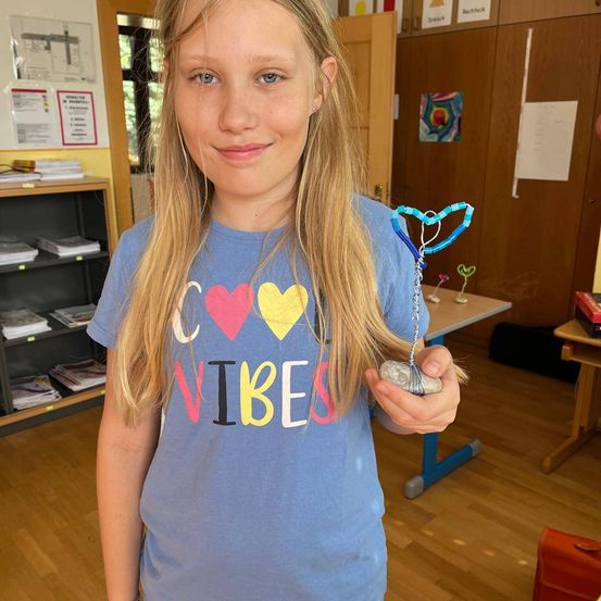 Ein Mädchen mit blonden Haaren hält einen Stein mit einer blauen Schnur. Sie trägt ein blaues T-Shirt mit dem Wort 'Vibes' darauf. Hinter ihr steht ein Holzschrank mit Büchern und ein Tisch.