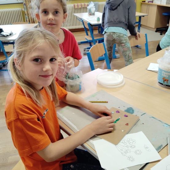 Ein Mädchen in einem orangefarbenen Shirt sitzt an einem Tisch in einem Klassenzimmer und arbeitet an einem Bastelprojekt. Ein weiteres Mädchen in Rot steht hinter ihr. Der Tisch ist mit Bastelutensilien wie Ton und Papier bestückt.