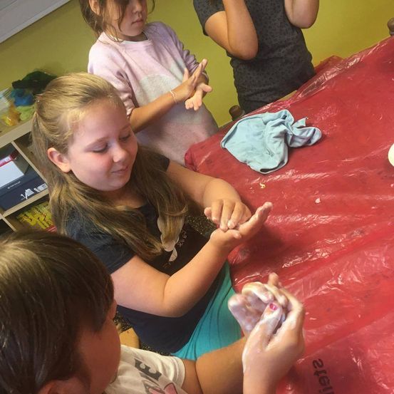 Drei Kinder sind drinnen; eine Mädchen lackiert ihre Nägel, während eine andere zuschaut. Eine dritte Mädchen steht hinter ihnen. Der Tisch ist mit einer roten Plastikfolie abgedeckt.