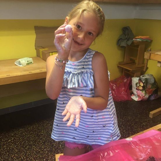 Ein kleines Mädchen steht in einem Raum, hält eine rosa Kugel aus Teig und lächelt, mit Mehl an den Händen. Hinter ihr ist eine gelbe Wand mit einer Kiste und einem Hocker.