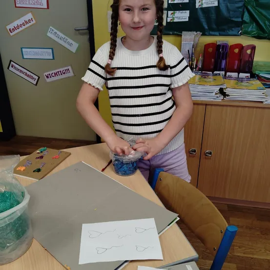 Ein junges Mädchen mit geflochtenen Haaren arbeitet in einem Klassenzimmer an einem Kunstprojekt. Sie trägt ein gestreiftes Shirt und lila Hose. Vor ihr liegt eine Plastiktüte mit blauem Glitzer und ein Papier mit Herz-Designs. Hinter ihr befindet sich ein Klassenzimmer mit verschiedenen Gegenständen auf einem Schreibtisch.