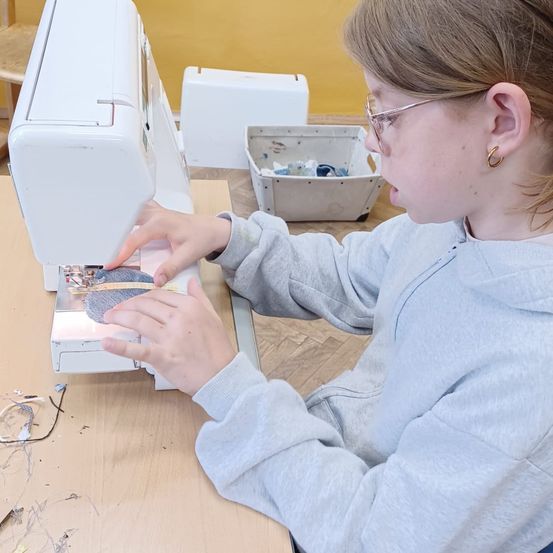 Ein Mädchen benutzt eine Nähmaschine, trägt eine Brille und ein blaues Sweatshirt. Sie konzentriert sich auf ihre Arbeit, mit Stoff und einer Aufbewahrungsbox in der Nähe.