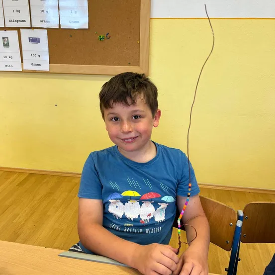 Ein Junge in einem blauen T-Shirt sitzt an einem Schreibtisch und hält einen langen Stock mit Perlen. Er lächelt. Hinter ihm ist eine Pinnwand mit Papieren und Gewichten.