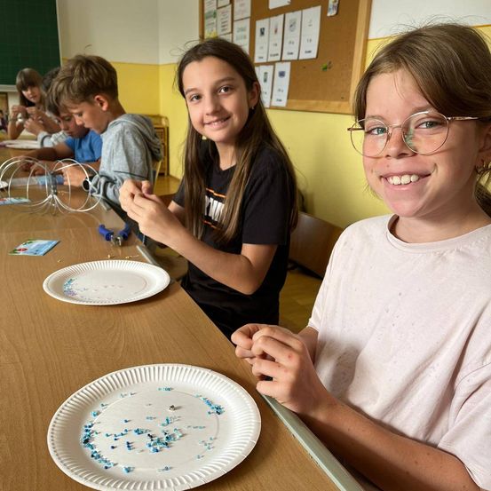 Schüler sitzen in einem Klassenzimmer an einem Tisch, zwei Mädchen konzentrieren sich auf ihr Projekt mit Tellern voller Perlen vor ihnen.