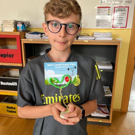 Ein junger Junge mit Brille und einem grauen Shirt mit 'Emirates' darauf hält eine Medaille und eine Karte mit der Aufschrift 'Mein Lesefitness-Pass'. Er steht in einem Raum mit Holzböden und einer gelben Wand. Hinter ihm steht ein Regal mit Büchern und Papieren und eine rote Box mit dem Wort 'Papier' darauf.