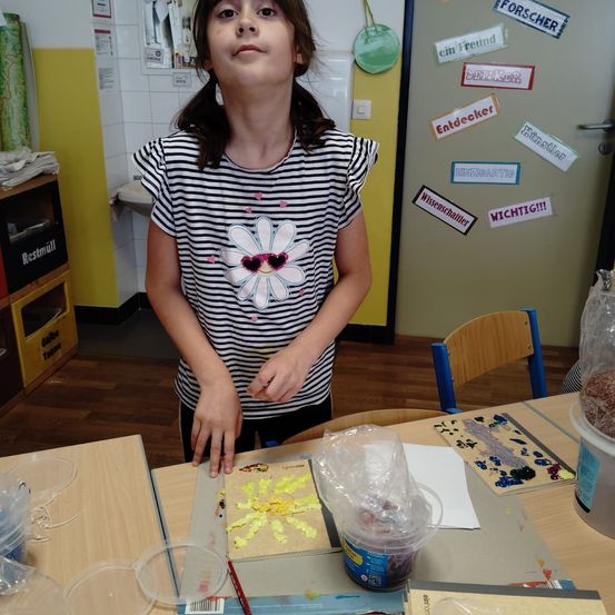 Ein Mädchen steht in einem Klassenzimmer vor einem Schreibtisch, der vor ihr steht, und hält ein Stück Papier mit Blumendesign in der Hand. Hinter ihr ist eine Tür mit verschiedenen Aufklebern zu sehen. Der Schreibtisch ist mit Gegenständen wie einem Plastikbehälter, einem Papiersack und einem Pinsel übersät.