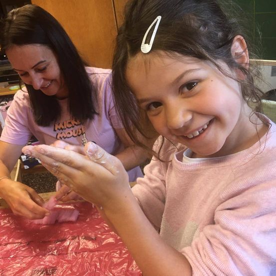 Ein lächelndes junges Mädchen benutzt ihre Hände, um mit einer Frau, die ein Micky-Maus-T-Shirt trägt, einen Bastelartikel herzustellen. Beide tragen rosa Langarmshirts.