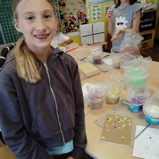 Ein junges Mädchen mit Zahnspange lächelt in einem Klassenzimmer, umgeben von Bastelmaterialien und einem Tisch.