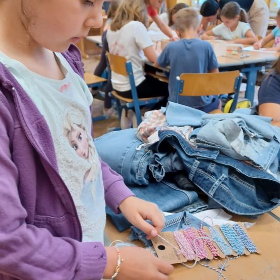 Ein junges Mädchen in einer lila Jacke arbeitet an Nähprojekten an einem Tisch, mit anderen Kindern in einem Klassenzimmer.