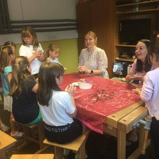 Eine Gruppe junger Mädchen versammelt sich um einen Tisch mit rotem Bezug. Zwei Frauen stehen da, eine hält eine Schüssel und die andere spricht mit den Mädchen.