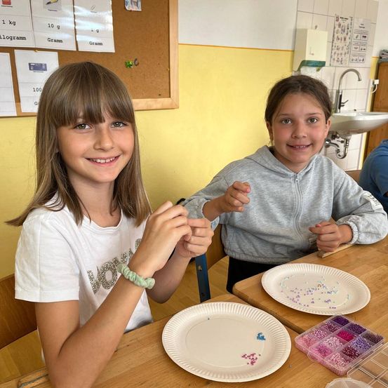 Zwei Mädchen sitzen lächelnd am Tisch und tragen Armbänder. Eines trägt ein weißes T-Shirt und das andere eine graue Kapuzenjacke. Vor ihnen stehen Pappteller mit Perlen. Dahinter ist eine Pinnwand und ein Waschbecken.