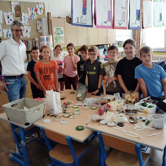 Eine Gruppe von Kindern und ein Mann stehen in einem Klassenzimmer. Sie versammeln sich um einen Tisch mit verschiedenen Gegenständen darauf, wie Knöpfe, Plastik und andere Objekte.