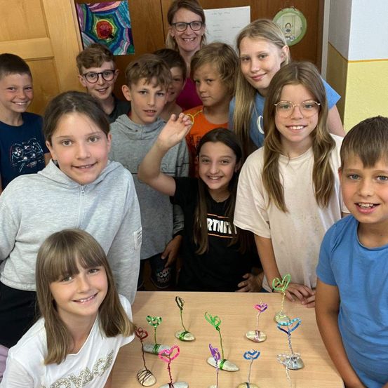 Eine Gruppe von Kindern steht um einen Tisch mit bunten Kunstwerken. Einige der Kinder tragen eine Brille und lächeln. Eine Frau steht hinter ihnen.