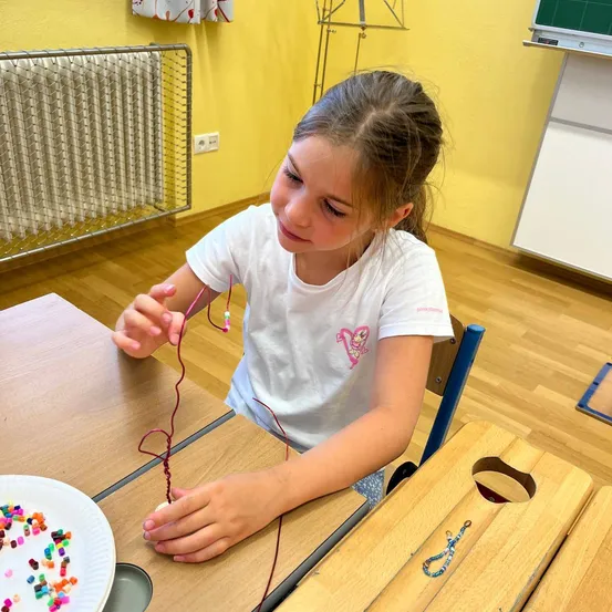 Ein kleines Mädchen sitzt an einem Schreibtisch und arbeitet an einem Perlenprojekt. Sie ist umgeben von hölzernen Tischen und Stühlen, und einer gelben Wand mit einem Heizkörper hinter ihr.