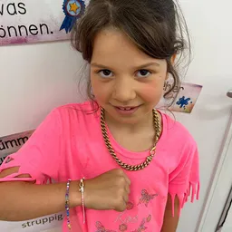 Ein junges Mädchen mit braunen Haaren, das ein rosa T-Shirt und eine goldene Halskette trägt, posiert für ein Foto mit einer weißen Wand im Hintergrund.