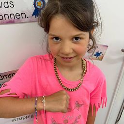 Ein junges Mädchen mit braunen Haaren, das ein rosa T-Shirt und eine goldene Halskette trägt, posiert für ein Foto mit einer weißen Wand im Hintergrund.